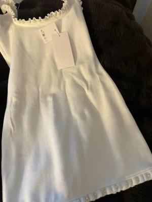 Vit kort klänning med pärldetaljer Zara - Supersöt vit kort klänning från Zara med volangkant och pärldetaljer vid halsringning och nederkant. Klänningen har en rak passform och är ärmlös, perfekt för sommarens alla tillfällen. Materialet är riktigt bra och klänningen är inte genomskinligt, Storlek M men skulle passa en S också.