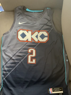 OKC Thunder basket tröja #2 - Snyggt basketlinne från med OKC Thunder-tryck och nummer 2, Gilgeous-Alexander. Linnet är mörkgrått med turkosa detaljer och geometriskt mönster på framsidan. Tillverkat i lätt och ventilerande material, perfekt för basket eller träning.