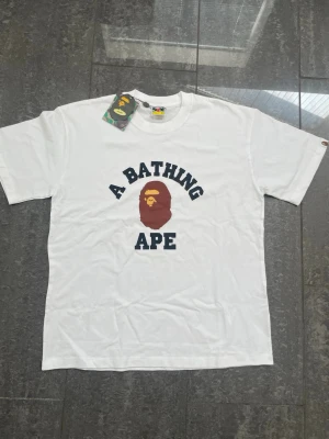 A Bathing Ape t-shirt - Helt ny vit t-shirt från A Bathing Ape med klassiskt tryck av apans ansikte och texten 'A BATHING APE' i svart på bröstet. T-shirten har rund hals och korta ärmar. Perfekt för dig som gillar streetwear och vill sticka ut med en ikonisk look.
