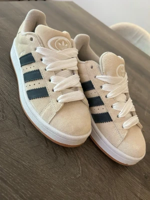 Beige Adidas Campus - Snygga sneakers från Adidas i beige mocka med klassiska svarta ränder på sidorna och vit sula. Skorna har breda vita skosnören och Adidas-logga på plösen. Perfekta för en stilren och sportig look. Campus helt nya endast testade. 
