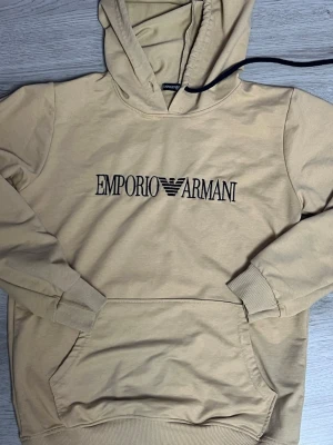 Beige hoodie från Emporio Armani - Snygg beige hoodie från Emporio Armani med svart broderad logga på bröstet. Klassisk känguruficka framtill, ribbade muddar och huva. Perfekt för en avslappnad och trendig stil. Materialet känns mjukt och bekvämt mot huden.