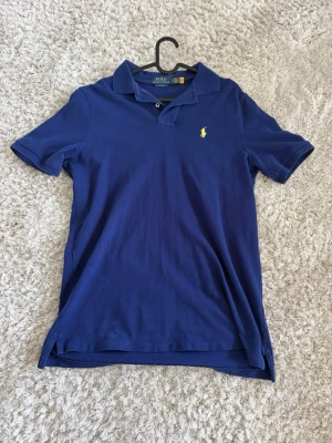 Blå pikétröja från Polo Ralph Lauren - Klassisk marinblå pikétröja från Polo Ralph Lauren med gul broderad logga på bröstet. Tröjan har krage, korta ärmar och knappar vid halsen. Tillverkad i mjuk bomull, perfekt till sommaren