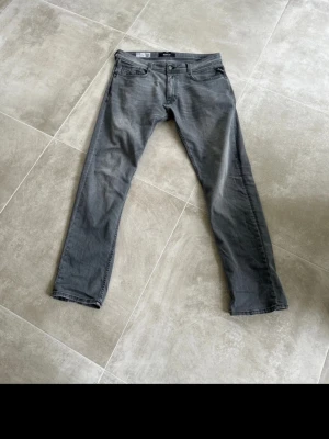Grå jeans från Replay - Snygga grå jeans från Replay med klassisk femficksmodell och raka ben. Jeansen har diskreta slitningar och detaljerad sömnad vid fickorna. Perfekta för en avslappnad och trendig look.