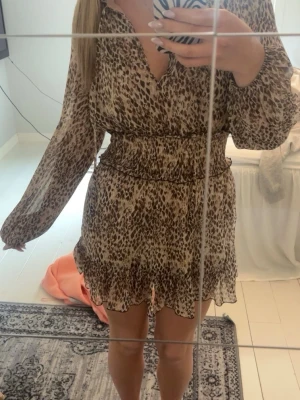 Leopardmönstrad kort klänning från Zara - Snygg kort klänning från Zara med leopardmönster i beige och bruna toner. Klänningen har v-ringning, långa ärmar och smockad midja som ger en fin passform. Volangdetalj nertill och lätt, luftig polyesterkvalitet.