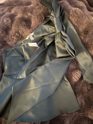 Grön satinsblus med oneshoulder - Snygg grön satinsblus från HM med oneshoulder-design och stor knytrosett vid axeln. Blusen har en glansig finish och elegant passform, perfekt för att sticka ut. Materialet är mjukt och följsamt satin.