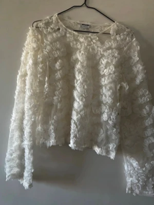 Vit blommig blus med struktur - Superfin vit blus från Monki med 3D-blommor över hela tyget. Blusen har rund halsringning och långa ärmar. Perfekt för dig som vill sticka ut med en unik och romantisk look. Materialet är lätt och transparent med blommiga detaljer.