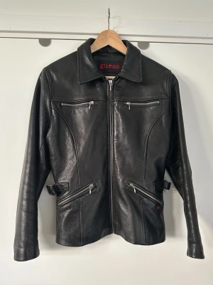 Svart skinnjacka från Guess, storlek S - Läcker svart skinnjacka från Guess i storlek S med dragkedja framtill. Cool modell med flera fickor med dragkedjor och spännen i sidorna för justering. En klassisk och tidlös kvalitetsjacka som passar till allt och kommer hålla säsong efter säsong. Nypris 8500:-. Mycket fint skick, som ny!