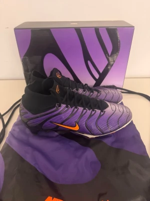 Nike lila fotbollsskor med orange swoosh - Säljer ett par Nike fotbollsskor i lila med svarta detaljer och orange swoosh. Skorna har strumpliknande skaft för extra stöd och är tillverkade i syntetmaterial med mönstrad ovandel. Perfekta för dig som vill sticka ut på planen med snygg design och bra grepp. Det bara att skicka om ni har frågor!!!