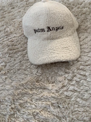 Vit teddykeps från Palm Angels - Säljer en vit keps från Palm Angels i mjukt teddy-material. Kepsen har böjd skärm och broderad logga framtill i svart. Justerbar rem baktill för perfekt passform. Perfekt accessoar för en trendig och avslappnad stil.