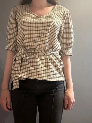 Rutigt blus med knytskärp - Snygg blus i beige och svart rutigt mönster med trekvartsärm och v-ringning. Blusen har ett knytskärp i midjan som ger en fin siluett och är tillverkad i ett lätt, strukturerat tyg. Perfekt för en trendig och avslappnad look.