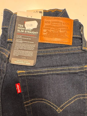 Levi's 724 High-Rise Slim Straight jeans - Mörkblå jeans från Levi's, modell 724 High-Rise Slim Straight med klassisk femficksdesign och gul kontrastsöm. Hög midja och rak, smal passform. Ikonisk läderpatch bak och röd Levi's-tagg. Stretchigt material för extra komfort.