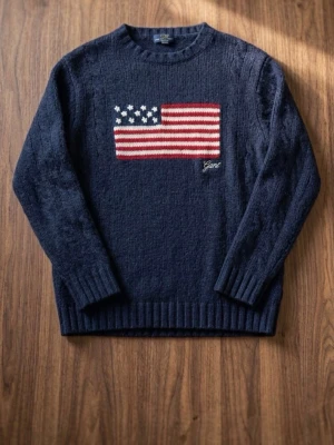 Gant-tröja med USA flagga - Snygg mörkblå stickad tröja från Gant med amerikansk flagga i rött, vitt och blått på bröstet. Tröjan har rund hals, ribbade muddar och Gant-logga broderad under flaggan. Perfekt för dig som gillar klassisk preppy-stil.
