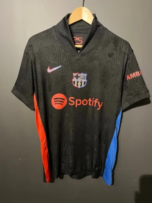 Svart FC Barcelona matchtröja Nike - Snygg svart FC Barcelona matchtröja från Nike med klubbmärke, röd Nike-logga och Spotify-tryck på bröstet. Röd och blå detalj längs sidorna, kort ärm och sportig krage. Är storlek L men skulle säga att den sitter mer som M.