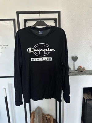 Svart långärmad Champion t-shirt - Svart långärmad t-shirt från Champion med stort vitt tryck på bröstet där det står 'Champion New York'. Klassisk rund halsringning och avslappnad passform. Perfekt för en streetwear-look.