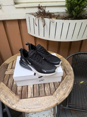 Nike Air Max 270  - Svarta Nike Air Max 270 sneakers med vit sula och synlig Air Max-dämpning i hälen. Skorna har en sportig design med mesh-överdel, svarta skosnören och små vita Nike-loggor på sidan och tån. Perfekta för dig som vill ha ett par bekväma skor nu till sommaren att kunna glida runt i. Passar även perfekt att ha på idrotten, gympa skor, springskor osv. Skick 10/10 inga skador alls! 