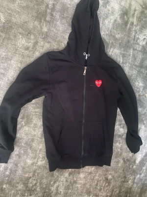 Heart and soul hoodie  - Svart hoodie med dragkedja och huva, samt en ficka framtill. På bröstet sitter ett rött hjärta med ögon, typiskt för Comme des Garçons Play. Perfekt för en avslappnad streetwear-look.