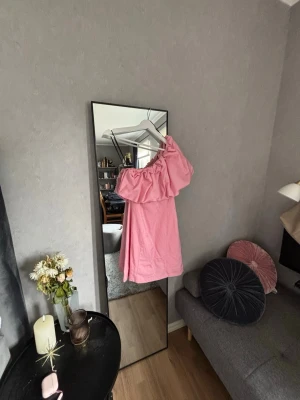 Rosa oneshoulder klänning - Säljer en rosa kortklänning med oneshoulder-design och volangdetalj över axeln. Klänningen har en figurnära passform och är perfekt för dig som vill sticka ut. Materialet känns mjukt och lätt, troligen bomull eller polyester, den har däremot några få fläckar precis under volangen (se bild 3) men som inte syns då armen täcker det. Supersnygg till sommaren!