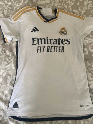 Real Madrid matchtröja Adidas 23/24 - Säljer en vit Real Madrid matchtröja från Adidas säsongen 23/24 med gulddetaljer på axlarna och mörkblå inslag. Tröjan har klubbmärke, Adidas-logga och Emirates Fly Better-tryck på bröstet. Tillverkad i lätt och ventilerande funktionsmaterial.