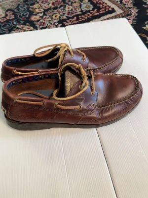 Bruna seglarskor från Timberland - Klassiska bruna seglarskor från Timberland i äkta läder med beige snörning och detaljer i mocka. Skorna har en robust sula och dekorativa sömmar runt tån. Perfekta för en avslappnad och stilren look.