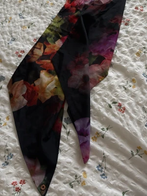 Svart scarf med blommigt mönster från ted baker - Halsduken är smal och långsmal i formen, perfekt att knyta runt halsen eller i håret. Materialet ser ut att vara mjukt och glansigt, troligen polyester eller siden.