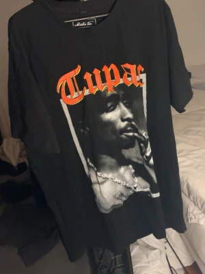 Svart Tupac t-shirt  - Cool svart t-shirt från Maldita med stort Tupac-tryck framtill i rött och gult. Oversized passform och klassisk rund hals. Perfekt för dig som gillar streetwear och vill sticka ut med en ikonisk look.