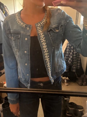 Ljusblå jeansjacka med pärldetaljer - Snygg ljusblå jeansjacka med pärlbroderi längs kragen och knäppningen. Jackan har två bröstfickor med lock och silverfärgade knappar. Kortare modell som passar perfekt till high waist jeans eller kjol. Perfekt för dig som vill sticka ut lite extra.