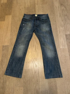 Blå bootcut jeans med slitningar från Denim & Co - Ett par blå bootcut jeans från Denim & Co med lätt slitna detaljer och klassisk femficksmodell. Jeansen har en något utsvängd passform nedtill och är tillverkade i denim. Perfekta för en avslappnad och trendig look.
