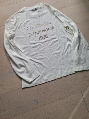 Vit långärmad topp Acne Studios med strass - Trendig vit långärmad topp från Acne Studios med texten 'Acne Studios STOCKHOLM 1996' framtill. Toppen har små dekorativa hål och är gjord i mjuk bomull. Perfekt för dig som gillar en edgy och avslappnad stil.