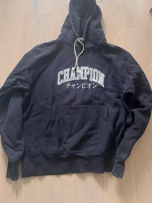 Mörk blå Champion hoodie med tryck - Mörk blå hoodie från Champion med stor vit logga och japansk text på bröstet. Klassisk känguruficka framtill, huva med vit dragsko och ribbade muddar. Mjuk och skön bomullsblandning, perfekt för en avslappnad stil.