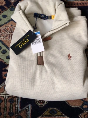 Beige halvzip-tröja från Polo Ralph Lauren - Snygg beige halvzip-tröja från Polo Ralph Lauren med klassisk broderad logga på bröstet. Tröjan har dragkedja med läderdetalj och är tillverkad i mjukt material. Perfekt för en stilren och avslappnad look.