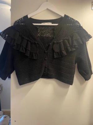 Svart kofta med volanger från Zara  - Snygg svart kofta från Zara med virkade detaljer och volanger över axlarna. Kortare modell med knappar framtill och trekvartsärmar. Perfekt för dig som gillar en trendig och unik look.