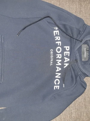 Mörkblå hoodie från Peak Performance - Snygg mörkblå hoodie från Peak Performance med vit logga framtill. Klassisk modell med huva och dragsko samt magficka. Mjuk insida och ribbade muddar vid ärmslut och nederkant. Perfekt för en avslappnad och sportig stil.