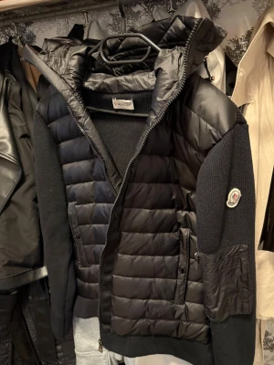Moncler Cardigan - Snygg svart pufferjacka från Moncler med stickade ärmar och huva. Jackan har quiltad front och rygg, dragkedja, fickor med dragkedja och Moncler-logga på ärmen. Perfekt för kalla vinterdagar och ger en sportig, trendig look.