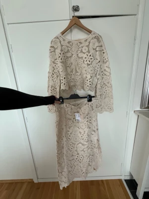 Beige virkade set med blommönster - Säljer ett superfint tvådelat set i beige virkad spets med blommönster. Setet består av en långärmad topp och en matchande kjol med vågig kant. Perfekt för dig som gillar boho-stil och unika detaljer. Materialet är mjukt och luftigt, vilket gör det bekvämt att bära. Kjol helt oanvänd men toppen har en liten fläck kolla sista bild.