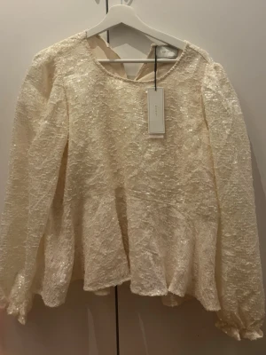 Glittrig krämvit blus från Neo Noir - Supersnygg krämvit blus från Neo Noir med glittrig, skimrande yta och lång ärm. Blusen har en lös passform och rund halsringning. Perfekt för dig som vill sticka ut lite extra med en trendig look.