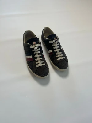 Moncler Monaco svarta sneakers med ränder storlek 40 - Svarta sneakers från Moncler med vita snören och klassiska Moncler-ränder i rött, vitt och blått på sidan. Skorna har låg profil, rund tå och ovandel i mocka. Moncler-logga syns på plösen och sidan. Perfekta för en stilren och sportig look. Skorna heter Moncler Monaco. De har storlek 40