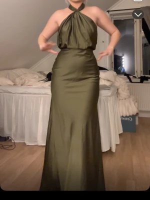 Olivgrön långklänning med halterneck - Snygg olivgrön långklänning med halterneck och figurnära passform. Klänningen har en glansig satin matrial och markerad midja. Perfekt för bal eller liknande! Nytt skick dock ingen prislapp kvar