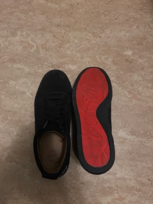 Svarta Louboutin sneakers med röd sula - Snygga svarta sneakers från Christian Louboutin med klassisk röd sula och silverbroderad logga på hälen. Skorna har lågt skaft, svart mocka och svarta skosnören. Perfekta för dig som vill sticka ut med exklusiv streetstyle.