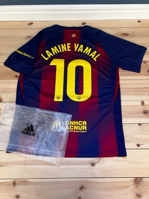 FC Barcelona matchtröja Lamine Yamal - Säljer en FC Barcelona matchtröja med Lamine Yamal och nummer 10 på ryggen. Tröjan är blå och vinröd med gula detaljer, klubbmärke och sponsorloggor på bröstet. Tillverkad i ett lätt och ventilerande material, perfekt för fotboll eller träning.