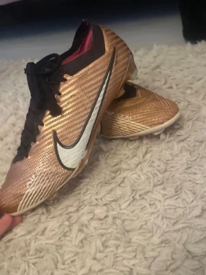 Guldiga Nike Mercurial fotbollsskor - Säljer ett par Nike Mercurial fotbollsskor i metallic guld med svart snörning och vit Nike-logga på sidan. Skorna har en smal passform och är tillverkade i syntetmaterial för bästa bollkontroll. Perfekta för dig som vill sticka ut på planen.