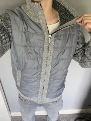 Grå cardiganjacka med stickade ärmar - Snygg grå cardiganjacka med stickade ärmar och quiltad front från Fashion BB. Jackan har dragkedja, hög krage och ribbade muddar. Perfekt för lager-på-lager och chill stil. Materialmix av stickat tyg och vadderad polyester framtill.