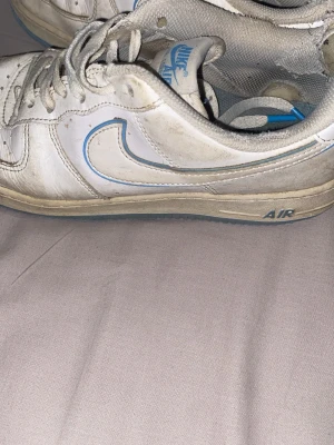 Nike Air Force 1 vita med blå detaljer - Nike Air Force 1 sneakers i vitt läder med blå detaljer och klassisk swoosh. Lågt skaft och snörning framtill. Sulan har AIR-tryck på sidan och insidan är vadderad för extra komfort. Perfekta för dig vill bara bli av med.