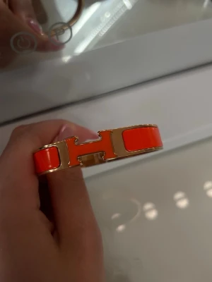 Orange armband  - Snyggt armband i orange emalj med guldfärgade metalldetaljer Stelt och elegant design som verkligen sticker ut och ger en lyxig känsla till din outfit.