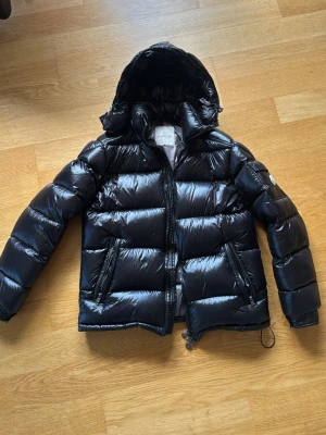 Svart dunjacka från Moncler med huva - Snygg svart dunjacka från Moncler med glansig finish och avtagbar huva. Jackan har två dragkedjefickor fram, Moncler-logga på ärmen och quiltad design. Perfekt för kalla vinterdagar och riktigt varm tack vare dunfyllningen.