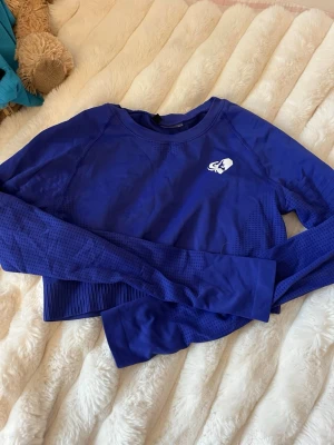 Blå långärmad crop top från Gymshark - Snygg blå långärmad crop top från Gymshark med vit logga på bröstet och ryggen. Tillverkad i stretchigt funktionsmaterial med ventilerande mesh-detaljer och ribbad nederkant. Perfekt för träning eller gymmet.