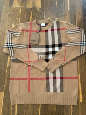 Beige stickad tröja Burberry - Snygg beige stickad tröja från Burberry med klassiskt rutmönster i svart, vitt, rött och brunt. Tröjan har rund halsringning och ribbade muddar vid ärmslut och nederkant. Perfekt för dig som gillar stilrena och trendiga plagg.