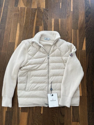  Moncler cardigan - Snygg beige cardigan från Moncler med stickade ärmar och hög krage. Västen har dragkedja framtill, två fickor och Moncler-logga på ärmen. Perfekt för lager-på-lager och riktigt trendig look. Materialmix av dun och stickat ger både värme och stil.