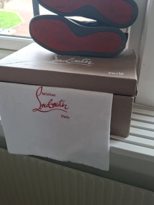 Christian Louboutin svarta sneakers - Svarta sneakers från Christian Louboutin med ikoniska röda sulor och svarta nitar på tån. Skorna har lågt skaft, snörning och är tillverkade i läder. Perfekta för dig som vill sticka ut med en edgy och lyxig stil.