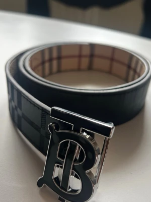 Burberry bälte - Stilrent bälte från Burberry med klassiskt rutigt mönster i svart och grått. Bältet har ett stort metallspänne med Burberrys ikoniska B-logga och insida i beige med rutor. Perfekt accessoar för att lyfta din outfit.