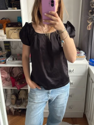 Svart puffärmad blus - Säljer en svart blus med puffärmar och rundad halsringning. Blusen har en lös passform och är tillverkad i ett glansigt material som ger en elegant känsla. Perfekt att styla med jeans eller kjol för en trendig look.